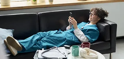 A woman in teal scrubs relaxes on a black leather sofa, smiling at her phone.